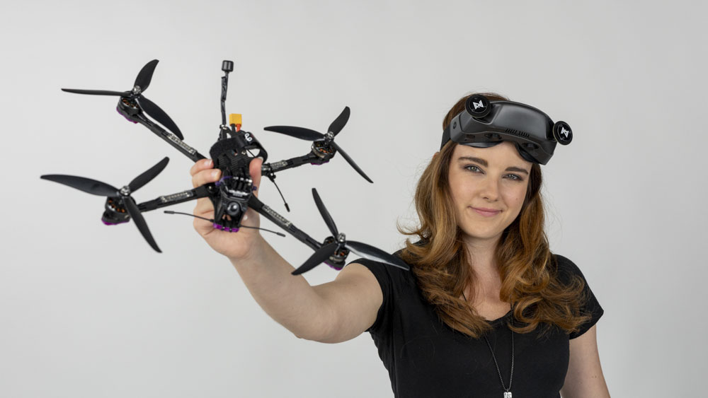 A woman with a confident smile holding a racing drone and wearing fpv (first person view) goggles on her forehead, shot on white photography backdrop.