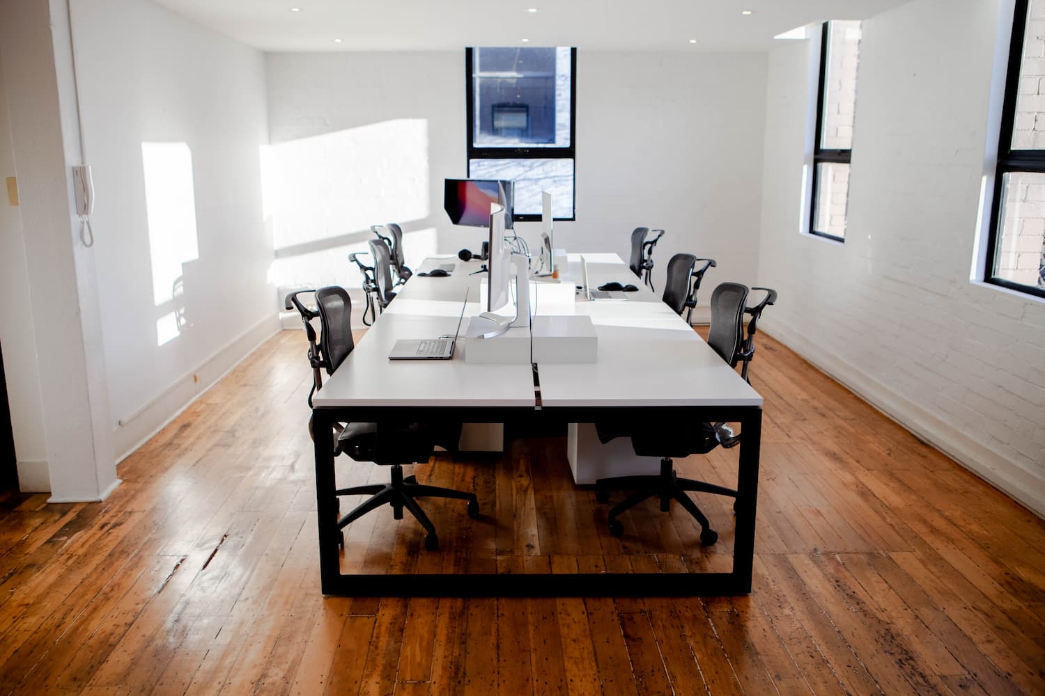 A modern office meeting room featuring a large white table with chairs around it, an iMac at one end, surrounded by white walls, large windows, and a hardwood floor.