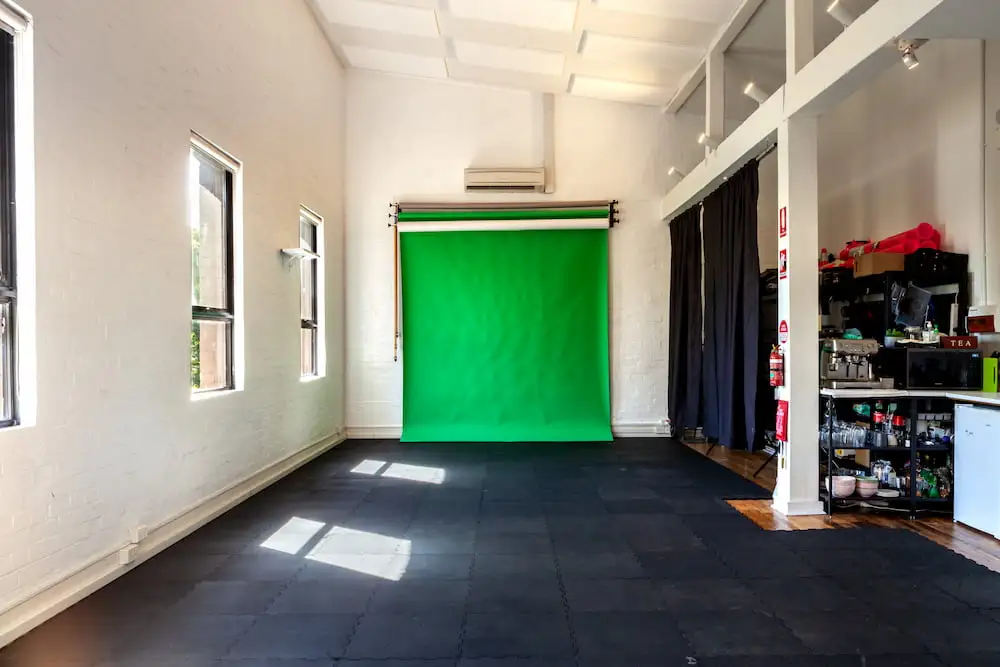 Green screen paper backdrop in a warehouse loft-style space, featuring natural light, white brick walls, and black rubber mats. Ideal for photography and video production.
