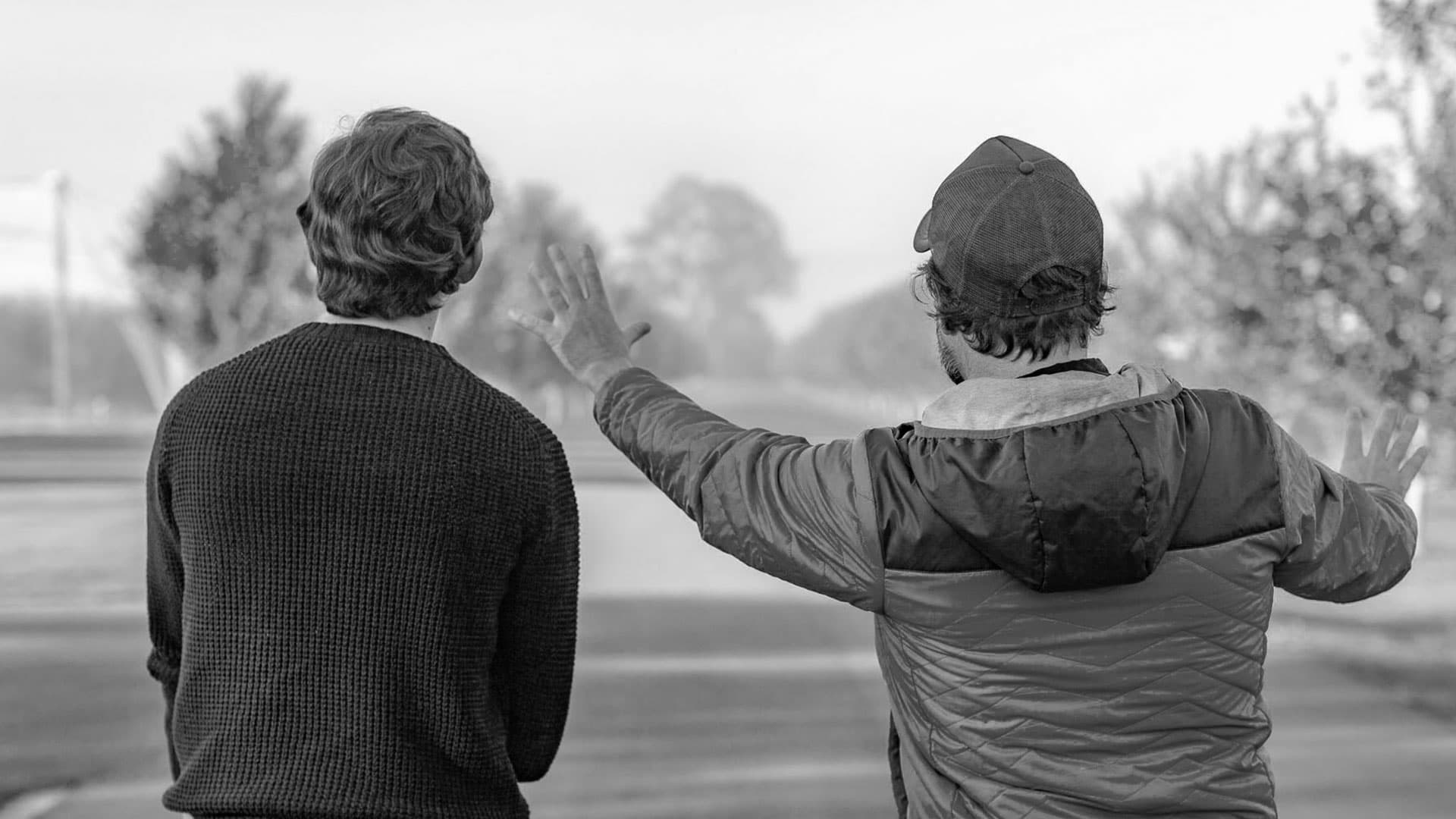 Two individuals engaged in a conversation, with one person gesturing expressively with their hand, possibly explaining something or telling a story, while standing outdoors with trees and an open space in the background. the image is captured in black and white, emphasizing the silhouettes and expressions of the subjects.
