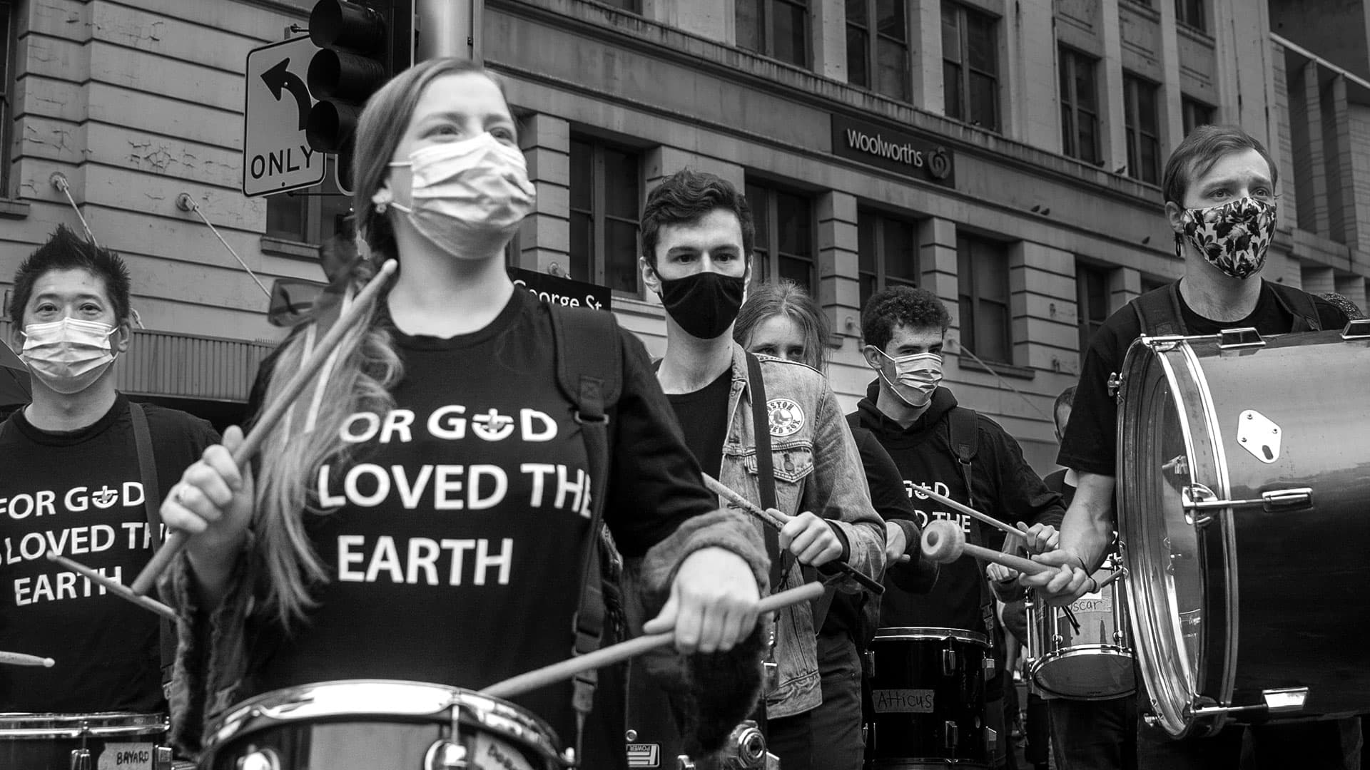 A group of activists wearing masks and playing drums during a street demonstration, united by a cause, with the prominent message on a participant's shirt expressing environmental concern.